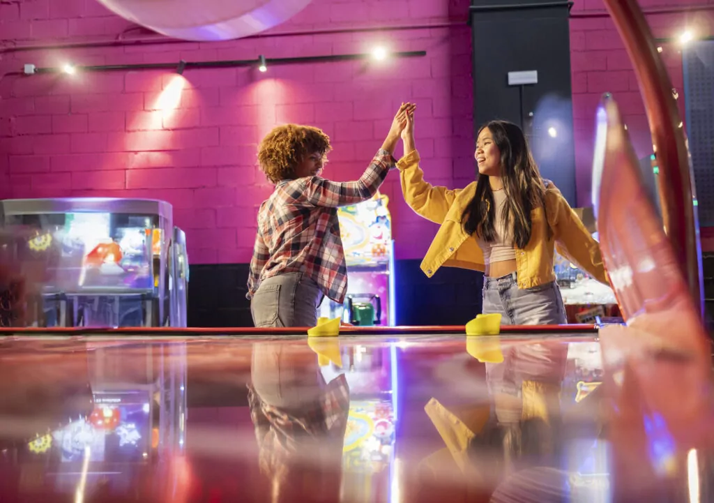 Two Friends Celebrating with High Five After Air Hockey Game. Two young women share a high five in an arcade after an exciting air hockey match, enjoying a fun moment together.