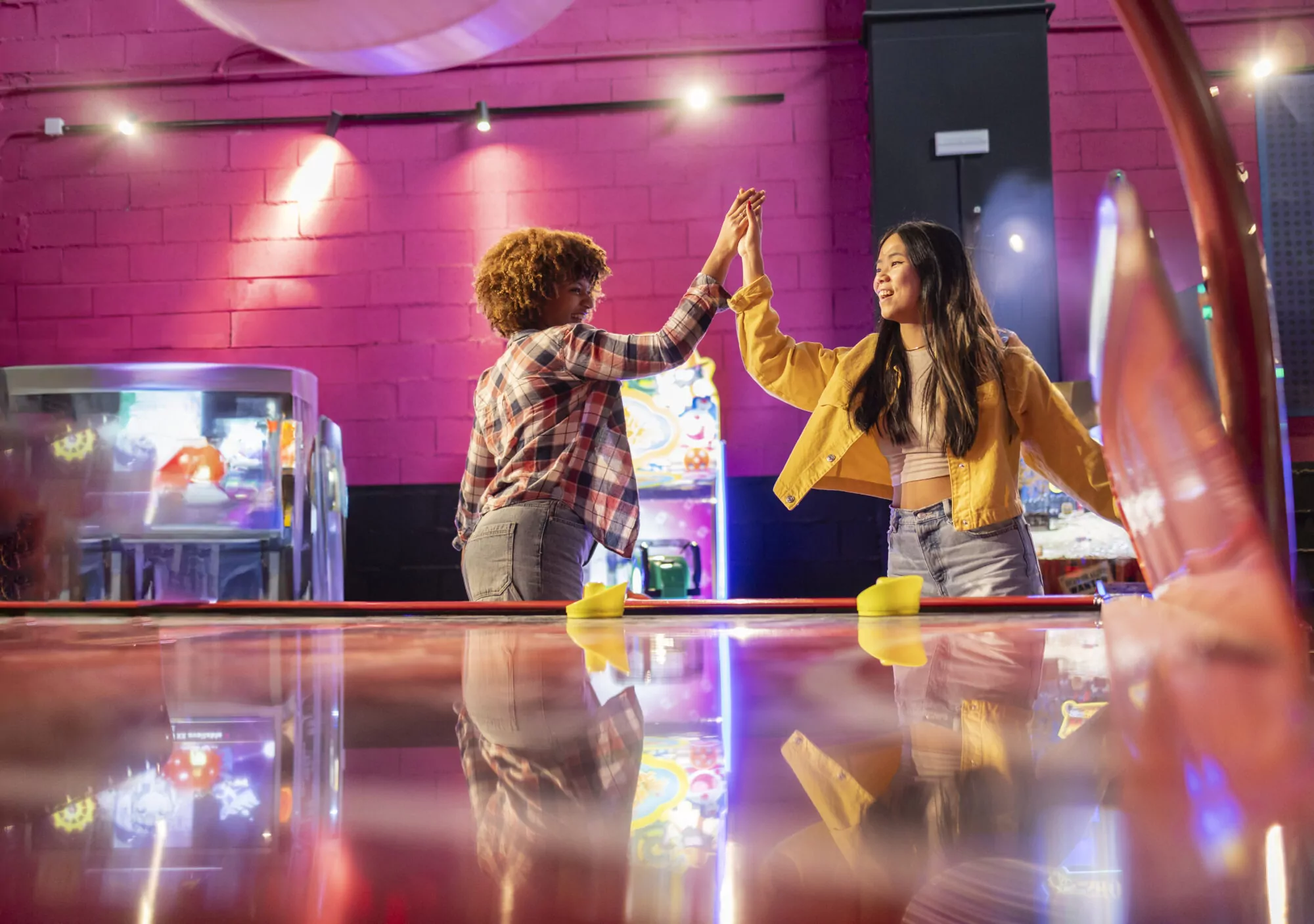 Two Friends Celebrating with High Five After Air Hockey Game. Two young women share a high five in an arcade after an exciting air hockey match, enjoying a fun moment together.